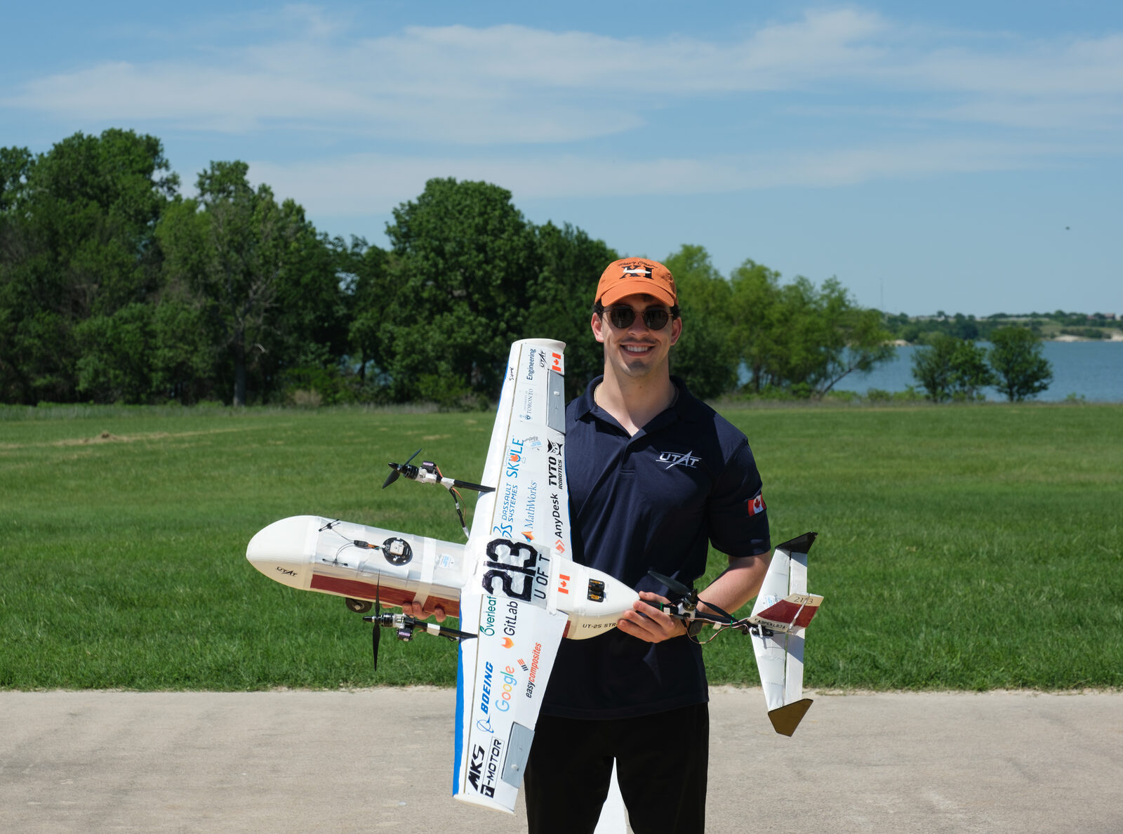The finished competition airframe held level at chest height on a grass field: white, two tractor motors on the wing, the number 213 on the wing and the fin, and UT-25 STRATUS lettered on the fuselage side.