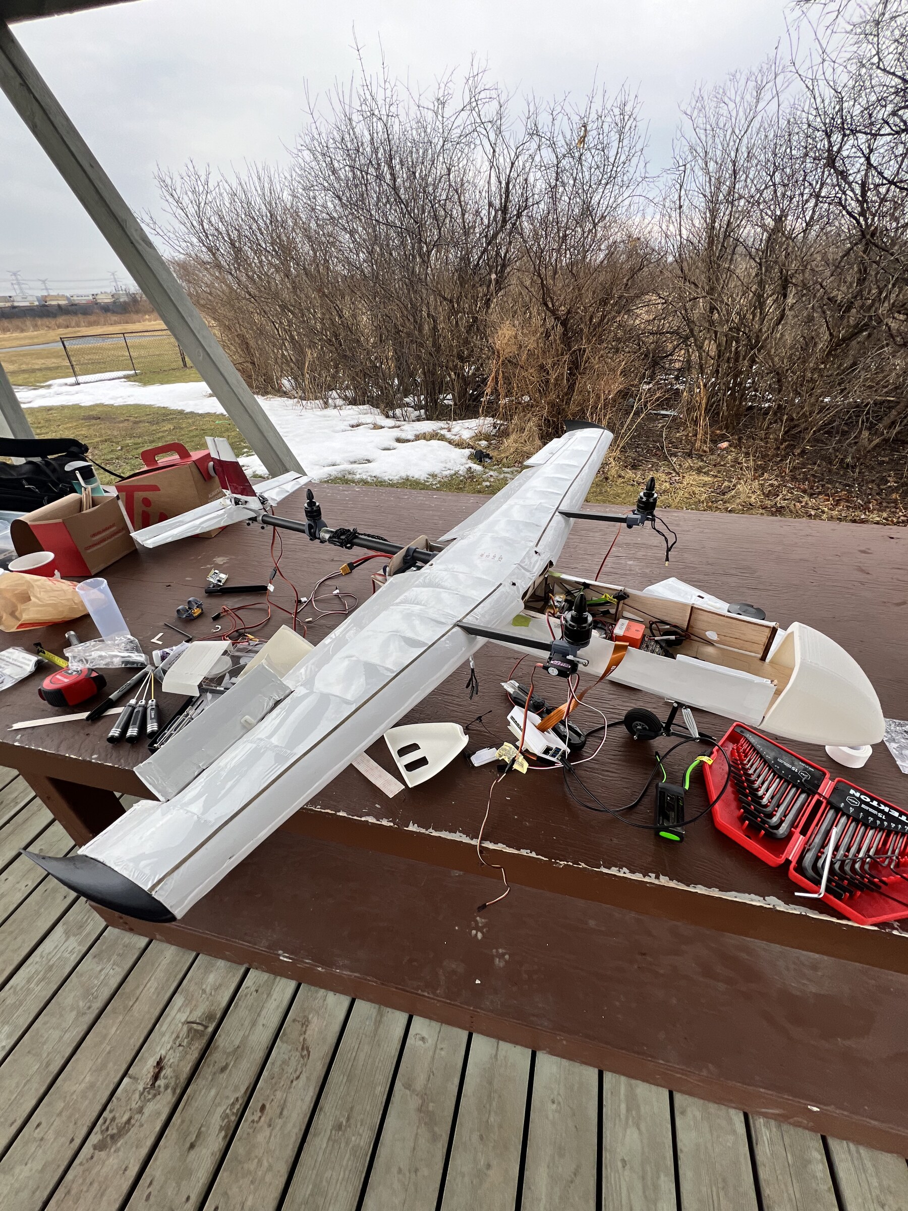 Stratus part-assembled on a picnic table at a flying field in late winter: wing joined and covered in white film with both motors mounted on the leading edge, fuselage open at the avionics and payload bay, nose cone, a mainwheel, a hex key set and a tape measure on the table beside it.