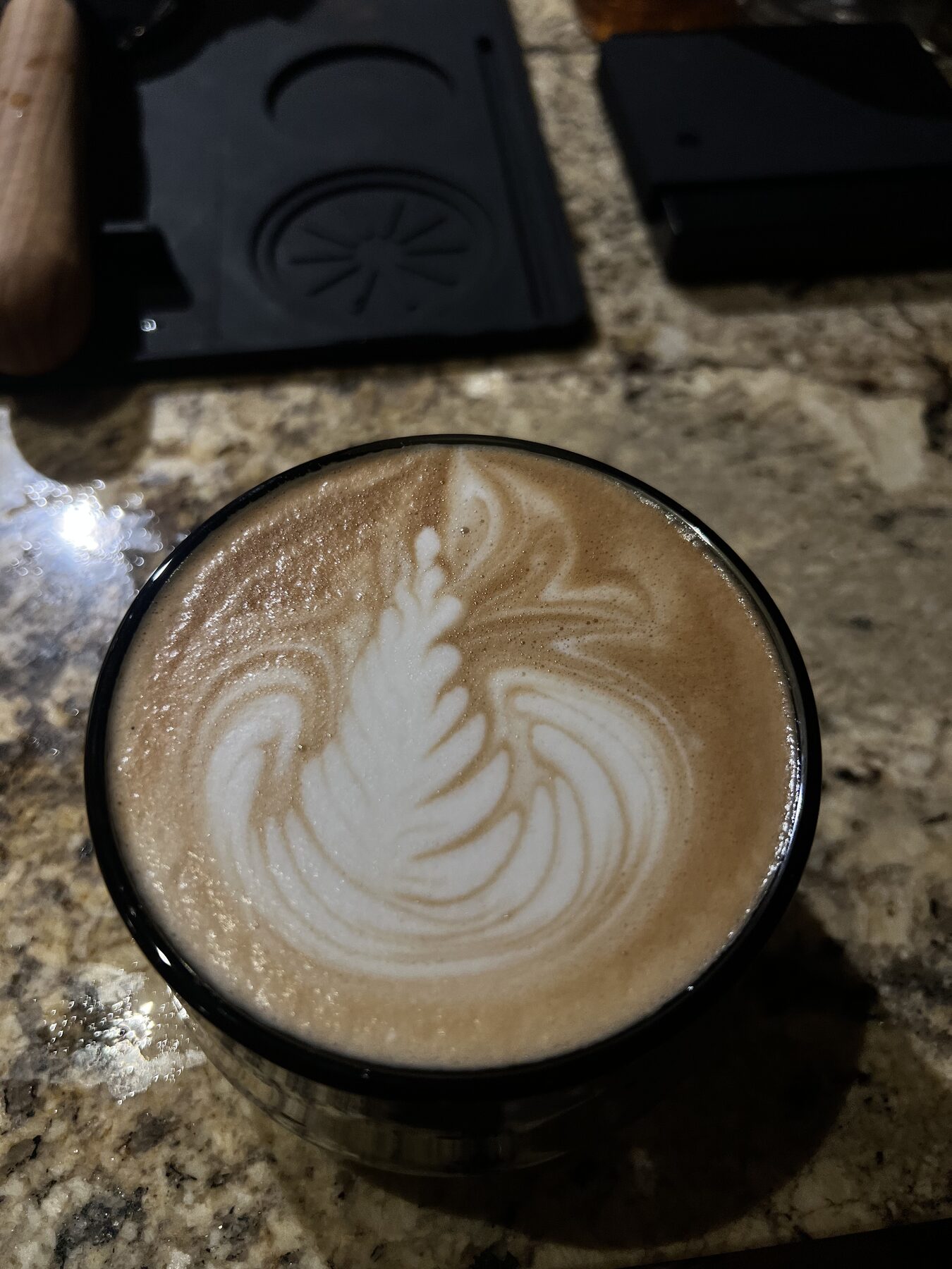Latte rosetta in a black-rimmed glass on granite, sharp leaf detail.