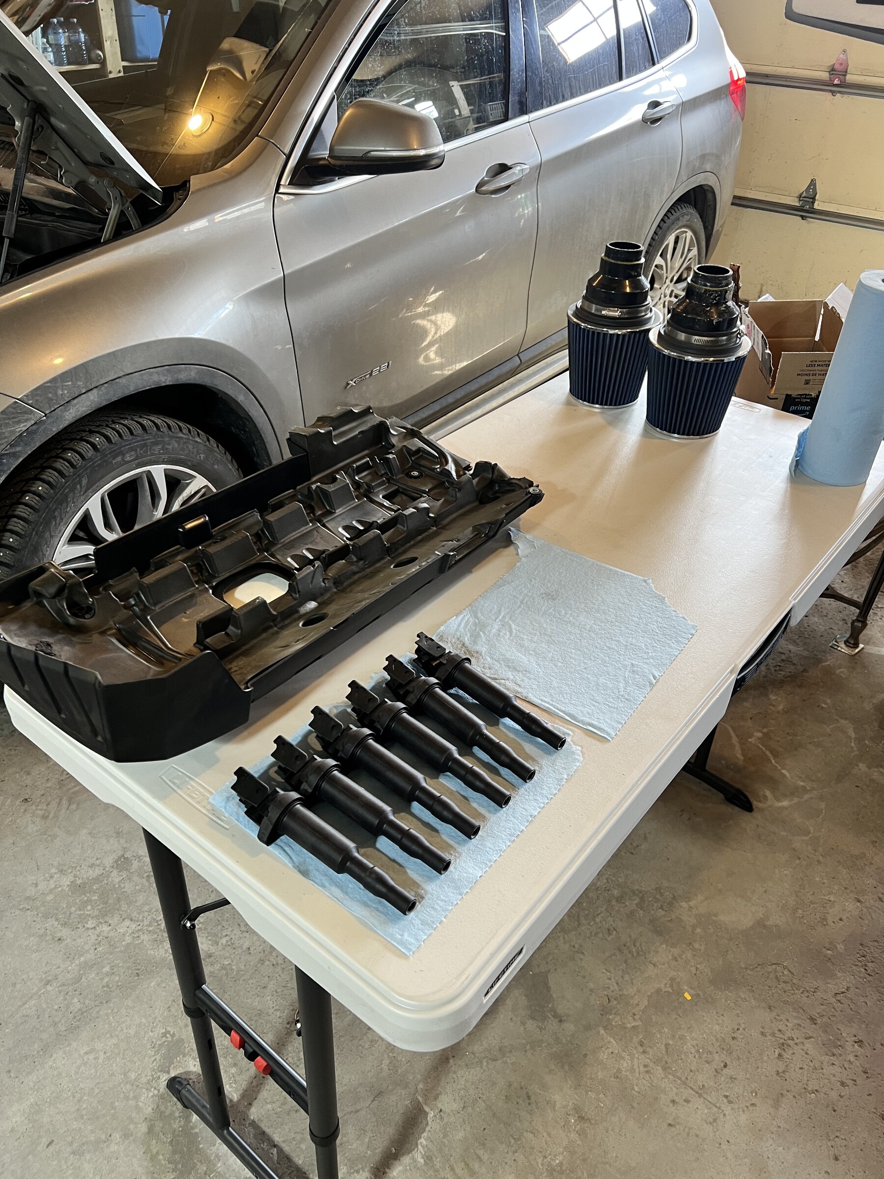 An engine cover and six ignition coils laid out in order on a folding table, two cone air filters beside them, and a car with its bonnet up behind.