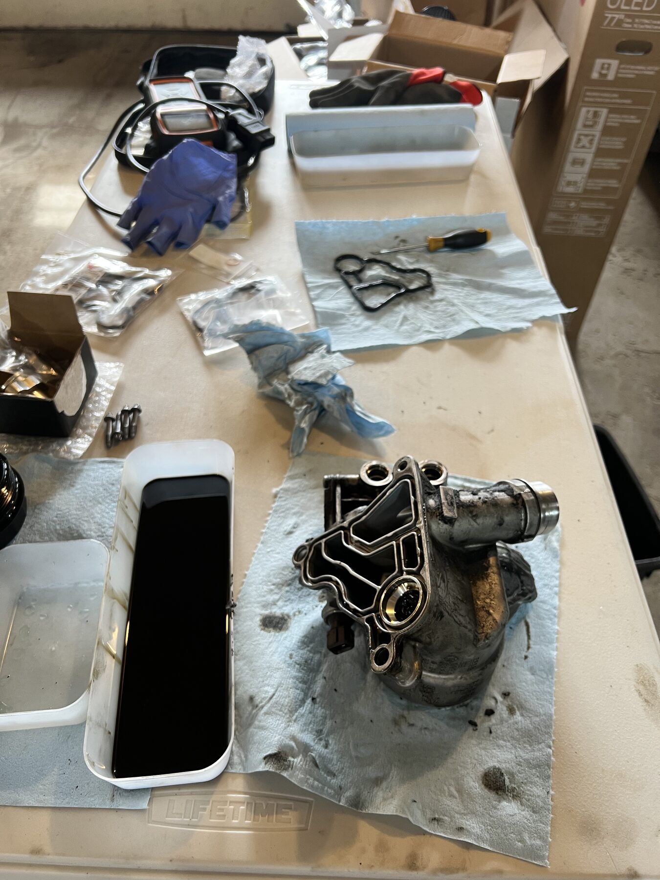 Oil filter housing pulled off the engine, on a fold-out workbench surrounded by gloves, gaskets, and tools.