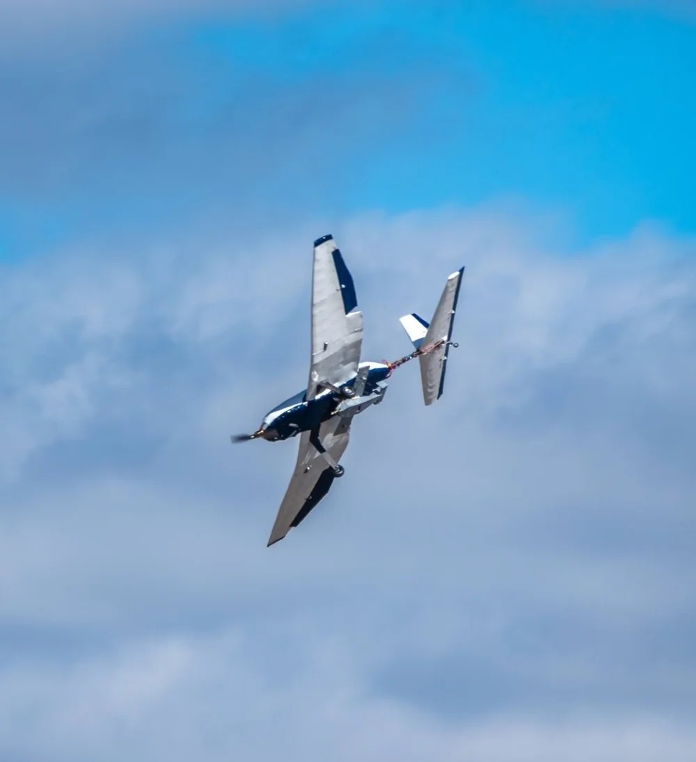 The same aircraft airborne against blue sky and broken cloud, banked hard enough that the wing is nearly edge-on to the camera, with the glider still carried on a slim boom beside the fuselage.
