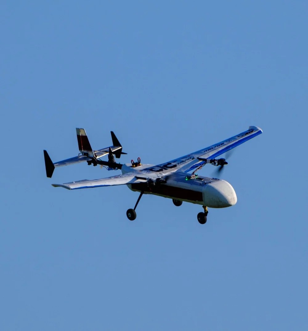 Stratus on another pass, seen from almost directly underneath: the full span of the wing across the frame with both motor mounts on it, the fuselage pod below, and the tail boom running back to a V-tail with a red and white marking.