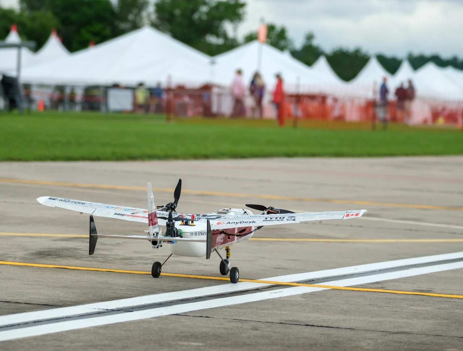 UT-25 Stratus standing on a taxiway: a white high-wing airframe with two motors mounted on the wing, a pod fuselage on fixed tricycle gear, sponsor decals across the wing, and competition marquees far out of focus behind it.