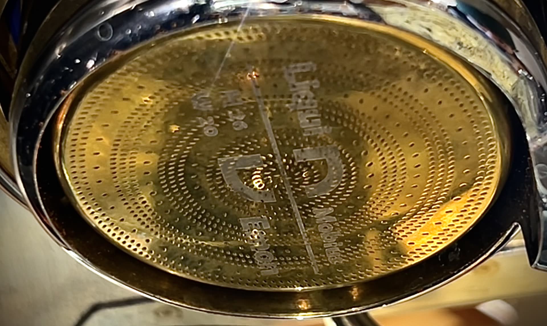 Looking up through the perforated dispersion screen of an espresso machine group head, brass and steel, with coffee above it.